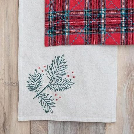 A wooden reindeer figurine sits on a red plaid cloth, which is placed on a light wood table with a white embroidered runner featuring pine sprigs and berries.