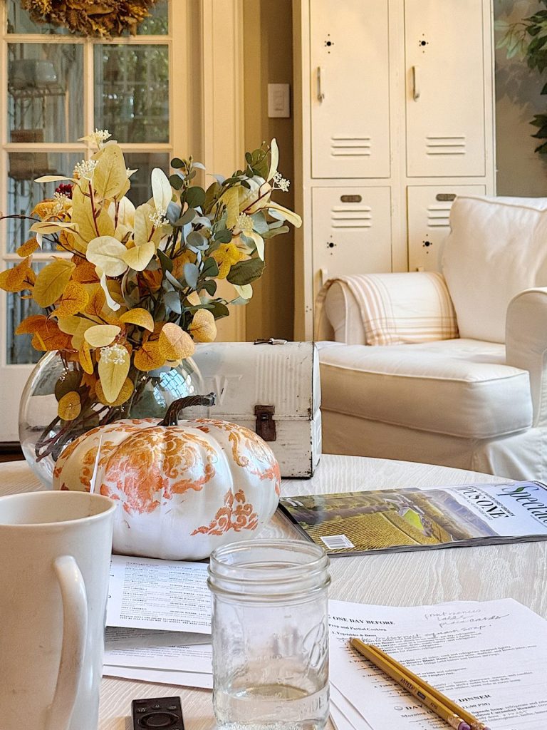 A glass vase with autumn leaves, a decorative pumpkin, magazines, papers, a mug, and a jar sit on a wooden table in a cozy, light-filled living room—perfect for moments of calm during a busy time.
