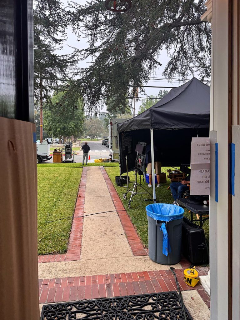 A view from a doorway shows a sidewalk leading to a street, with a black canopy tent, film equipment, and people working outside on a lawn.