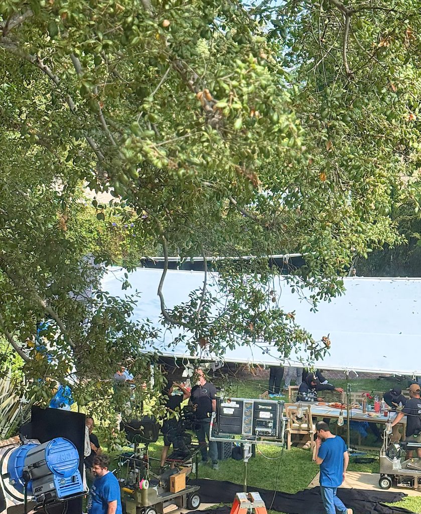 A film crew sets up equipment and workstations outdoors under trees and a large white screen, with various lights, monitors, and crew members visible.