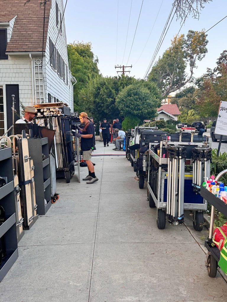 Film crew members stand on a residential sidewalk with equipment carts and gear, preparing for a shoot near a house and trees.