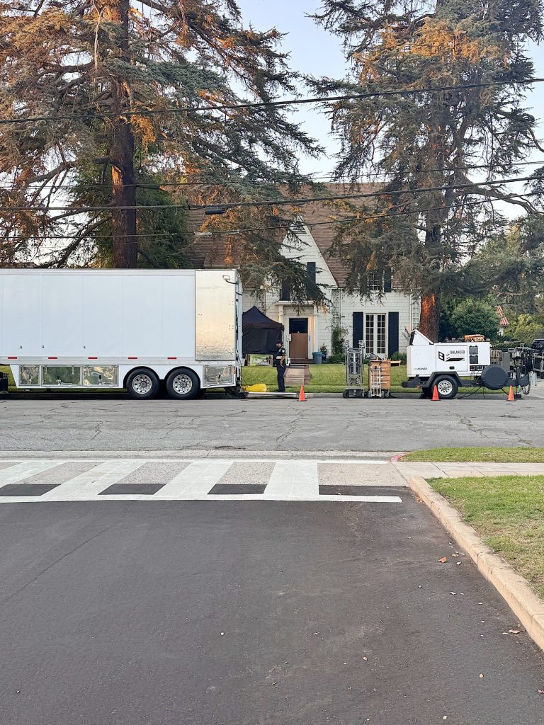 White film production truck and equipment trailer parked outside a house, with crew and gear visible on the front lawn, surrounded by tall trees.
