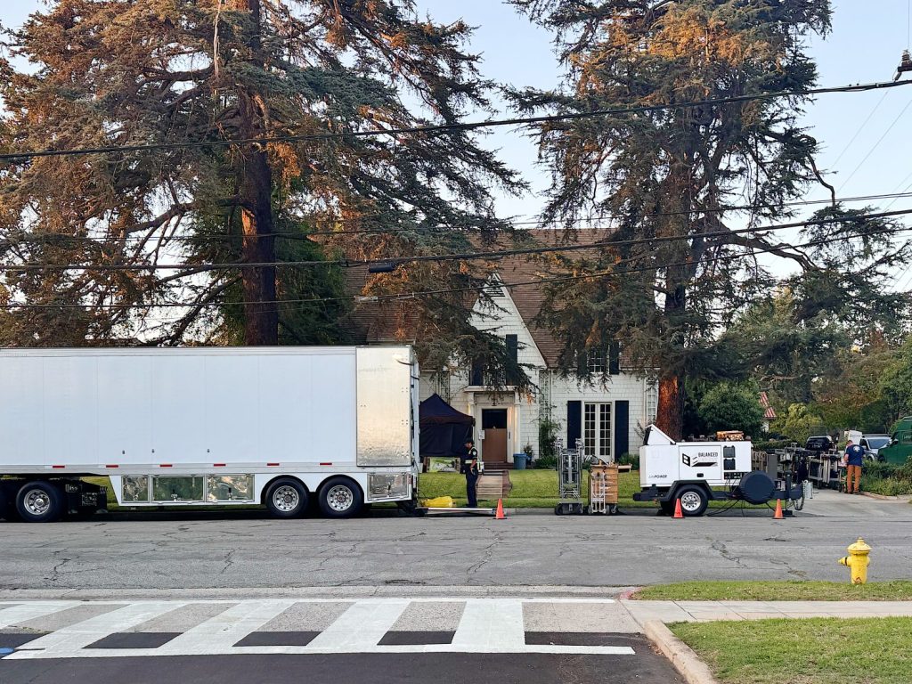 A large white truck and film equipment are set up in front of a house, indicating a film production taking place on a residential street.