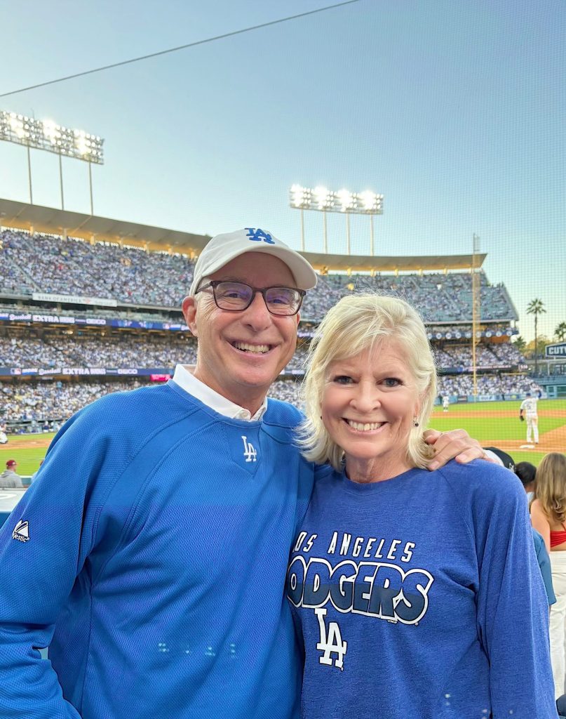 A man and woman wearing Los Angeles Dodgers apparel smile for a photo at a baseball stadium with a game in progress and a crowd in the stands behind them.