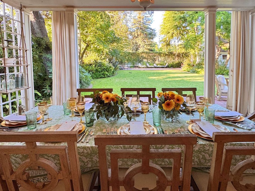 A dining table set with plates, glasses, and sunflower centerpieces invites a pause from a busy time, positioned by open doors facing a green backyard.