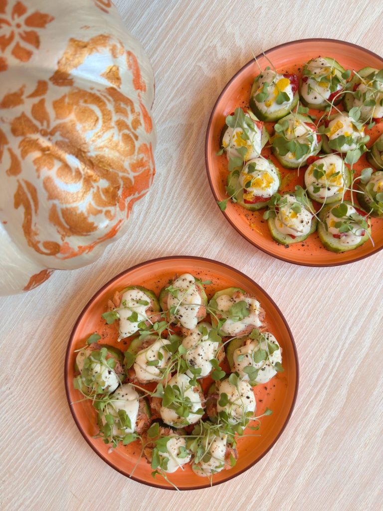 Two orange plates with bite-sized appetizers topped with herbs and creamy sauce sit on a light wooden table next to a decorative pumpkin, perfect for sharing during any busy time.