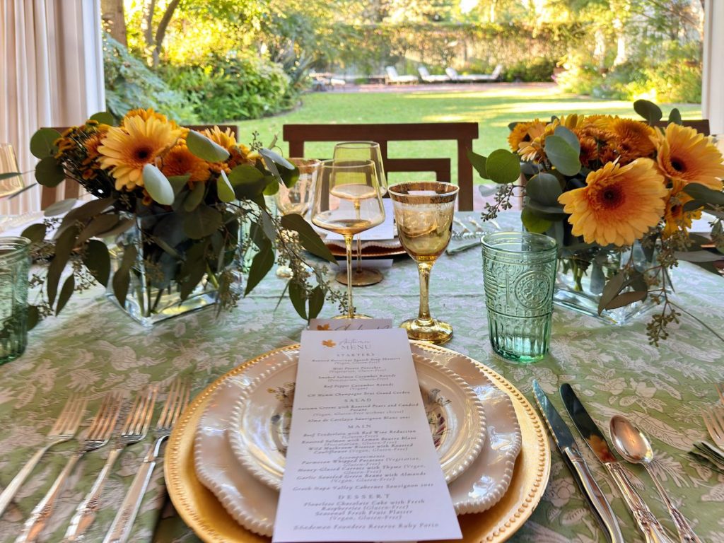 A formal table setting with floral centerpieces, glassware, and a printed menu on a stacked plate, set outdoors with a garden view—perfect for gathering during a busy time.