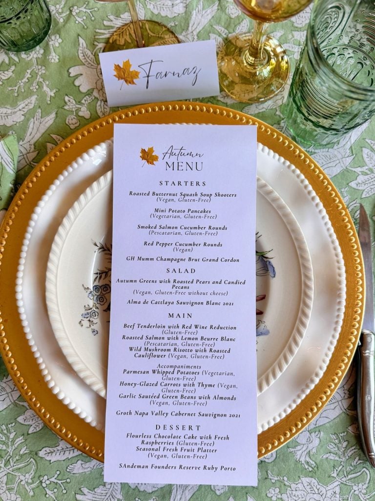 A formal autumn-themed dinner menu on a white plate with gold accents, surrounded by glassware and a place card reading "Farmaz" on a patterned tablecloth—perfect for a busy time of seasonal gatherings.