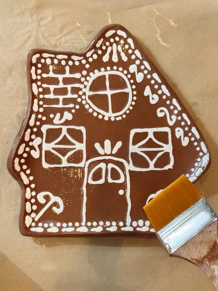 A decorated gingerbread house cookie with white icing details sits on parchment paper, next to a pastry brush.