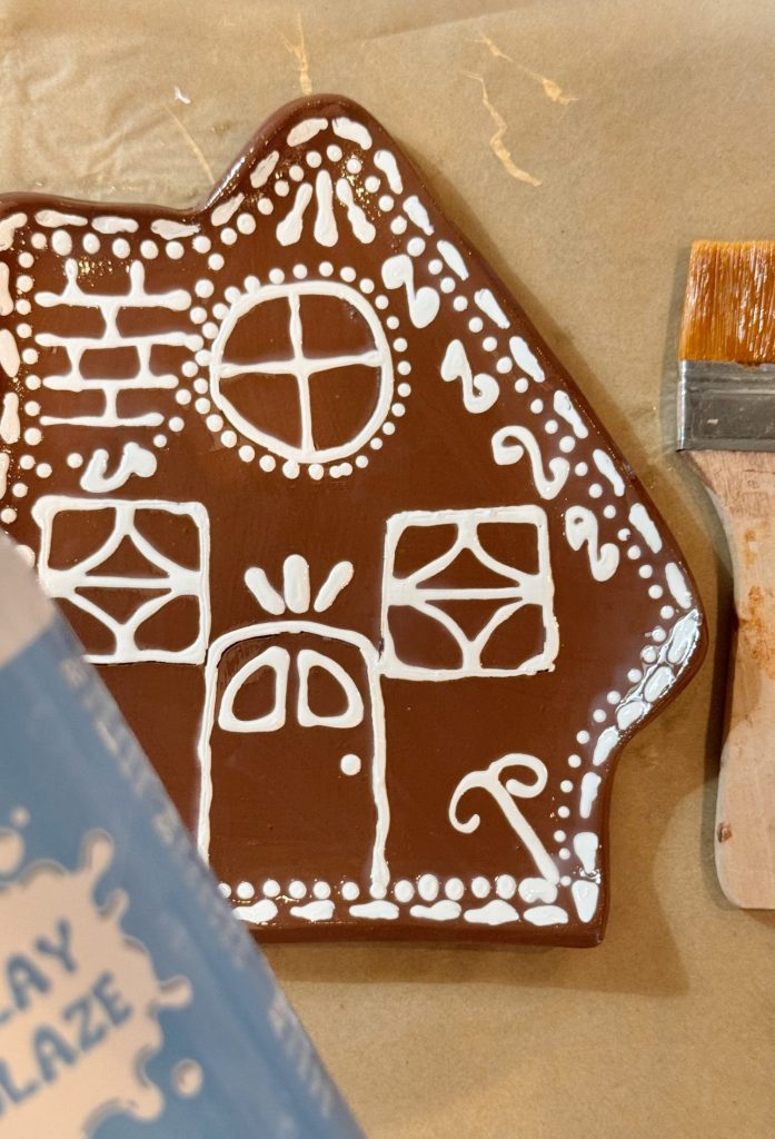 A brown ceramic piece shaped like a house is decorated with white icing-like patterns next to a paintbrush on parchment paper.