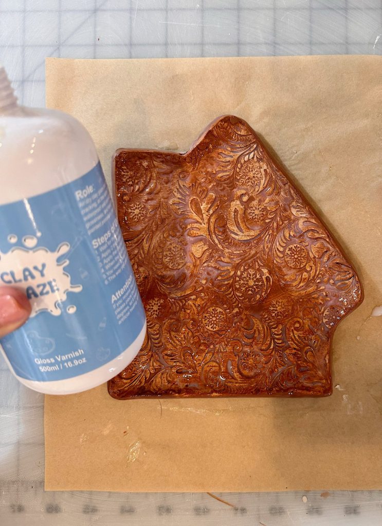 A hand holds a bottle of gloss varnish next to a brown, textured, house-shaped clay dish resting on parchment paper.