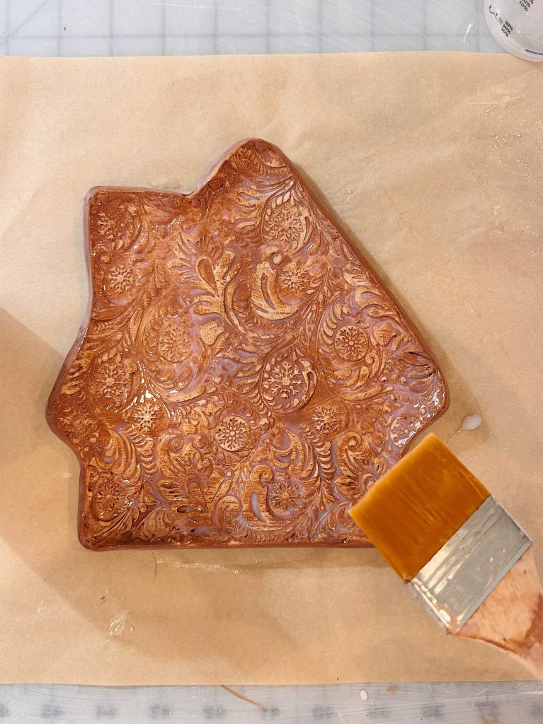 A decorative clay dish with an embossed floral pattern is being coated with a glossy finish using a broad paintbrush.