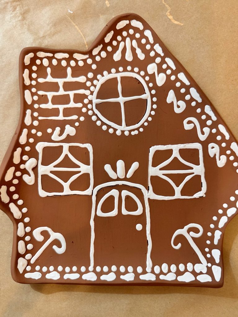 A gingerbread house-shaped cookie decorated with white icing patterns, featuring a door, windows, a circular window, candy canes, and dots.