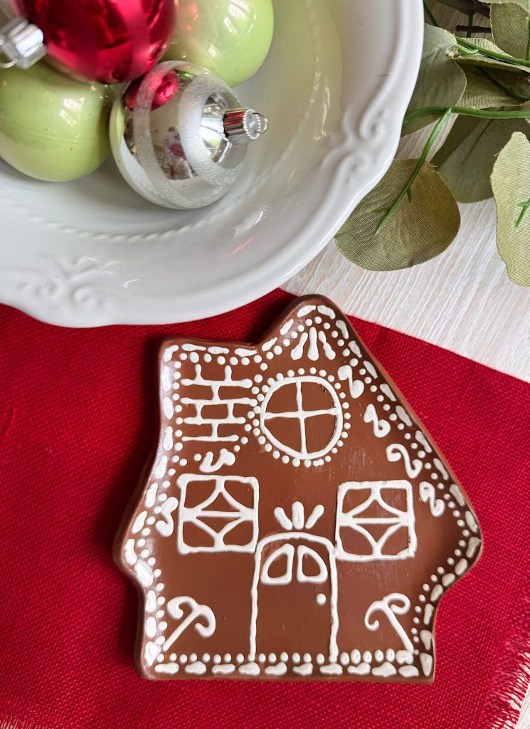 A gingerbread house cookie decorated with white icing sits on a red napkin next to a plate with Christmas ornaments and eucalyptus leaves.