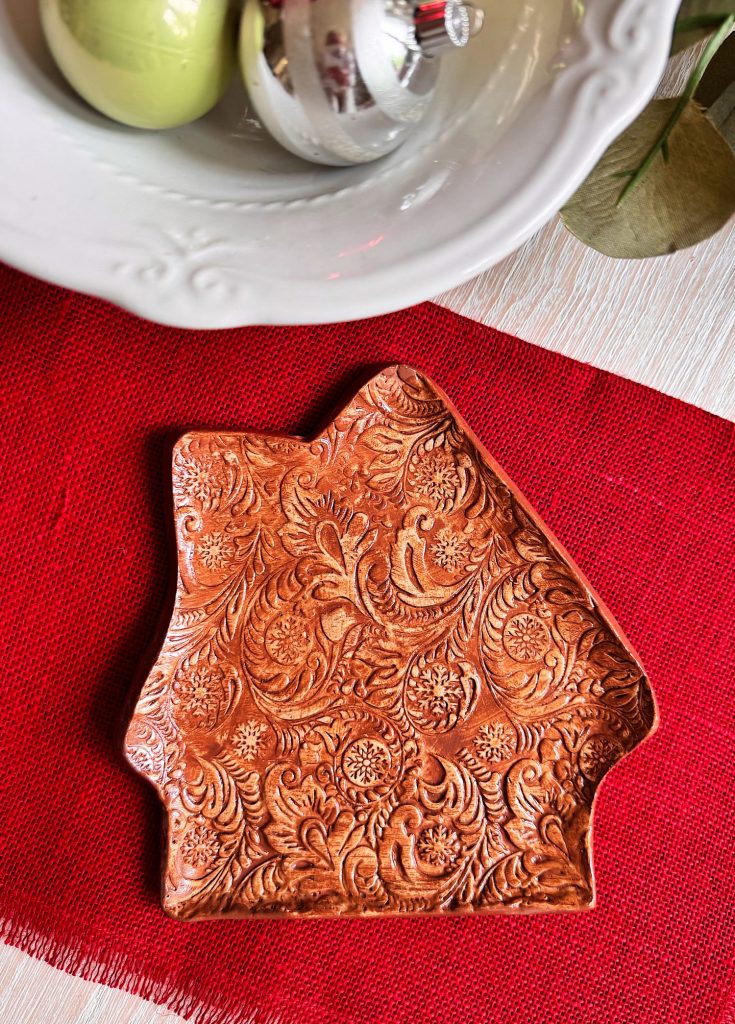 A decorative, brown ceramic dish with an embossed floral pattern, shaped like a house, placed on a red mat next to a white bowl containing ornaments.