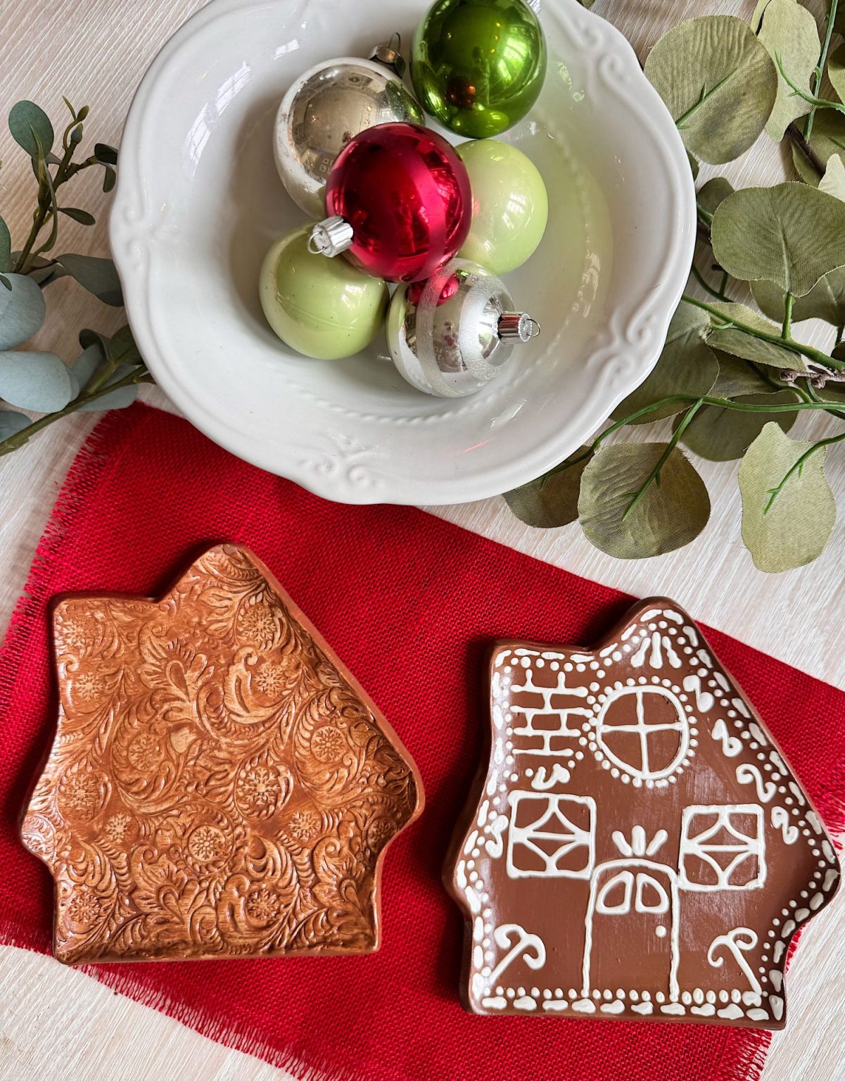 A white bowl holds green, red, and silver ornaments. Beside it are two gingerbread house-shaped ceramic dishes, one with patterns and one decorated as a house, on a red cloth.