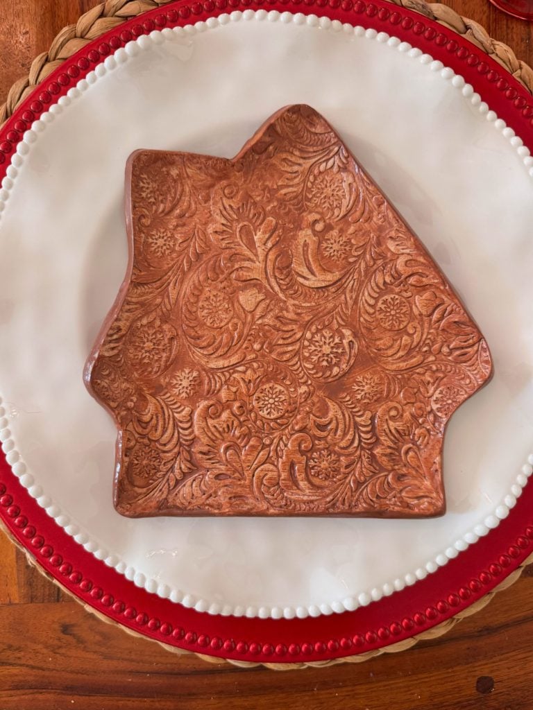 A brown, embossed ceramic plate in the shape of a house sits on a white plate with a red rim, placed on a woven placemat.