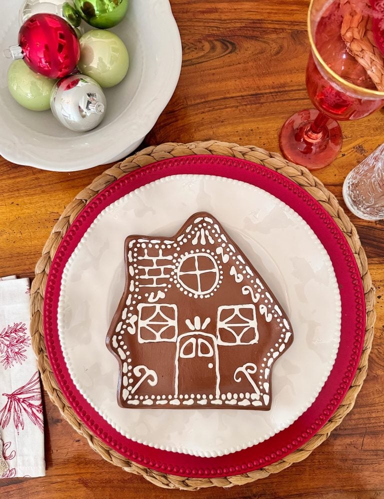 A gingerbread cookie decorated as a house is placed on a white plate with a red border, next to a bowl of Christmas ornaments and a red drink in a glass.