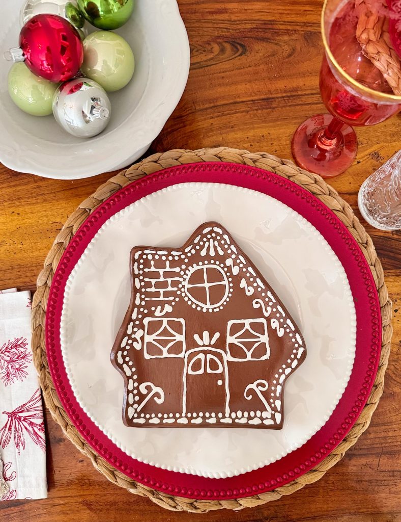 A gingerbread cookie decorated as a house is placed on a white plate with a red border, next to a bowl of Christmas ornaments and a red drink in a glass.