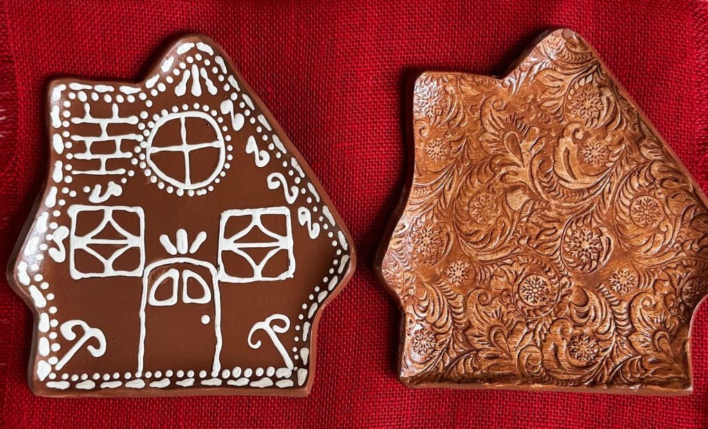 Two house-shaped ceramic plates on a red textured fabric; one has white icing-style decoration, the other has ornate embossed patterns.