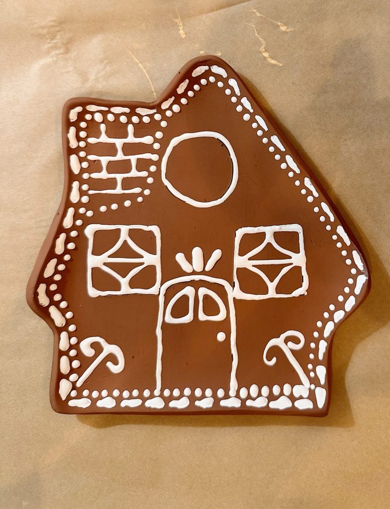 A gingerbread house cookie decorated with white icing, featuring a door, windows, a round window, and candy canes on a parchment paper background.