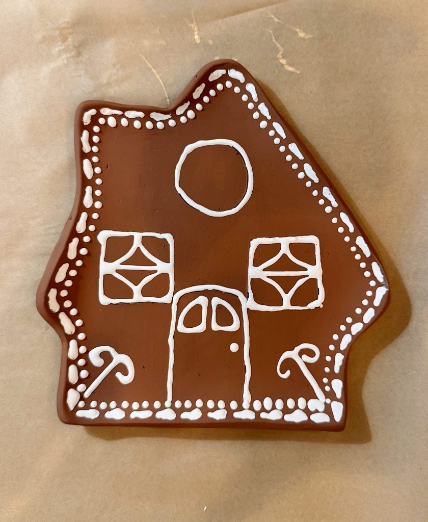 A gingerbread cookie shaped like a house, decorated with white icing to show windows, a door, a circle above, and two candy canes on either side of the door.