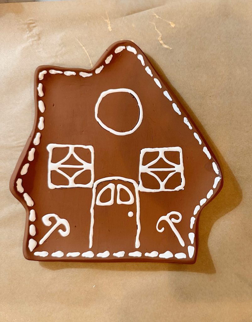 Brown gingerbread house cookie decorated with white icing outlines for windows, a door, and candy canes, placed on parchment paper.