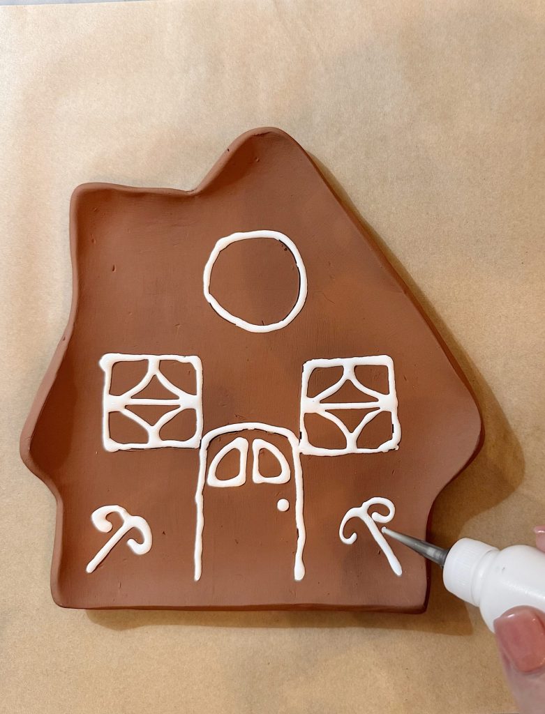 A hand uses white icing to decorate a brown, house-shaped cookie with windows, a door, a circle, and candy cane shapes on parchment paper.