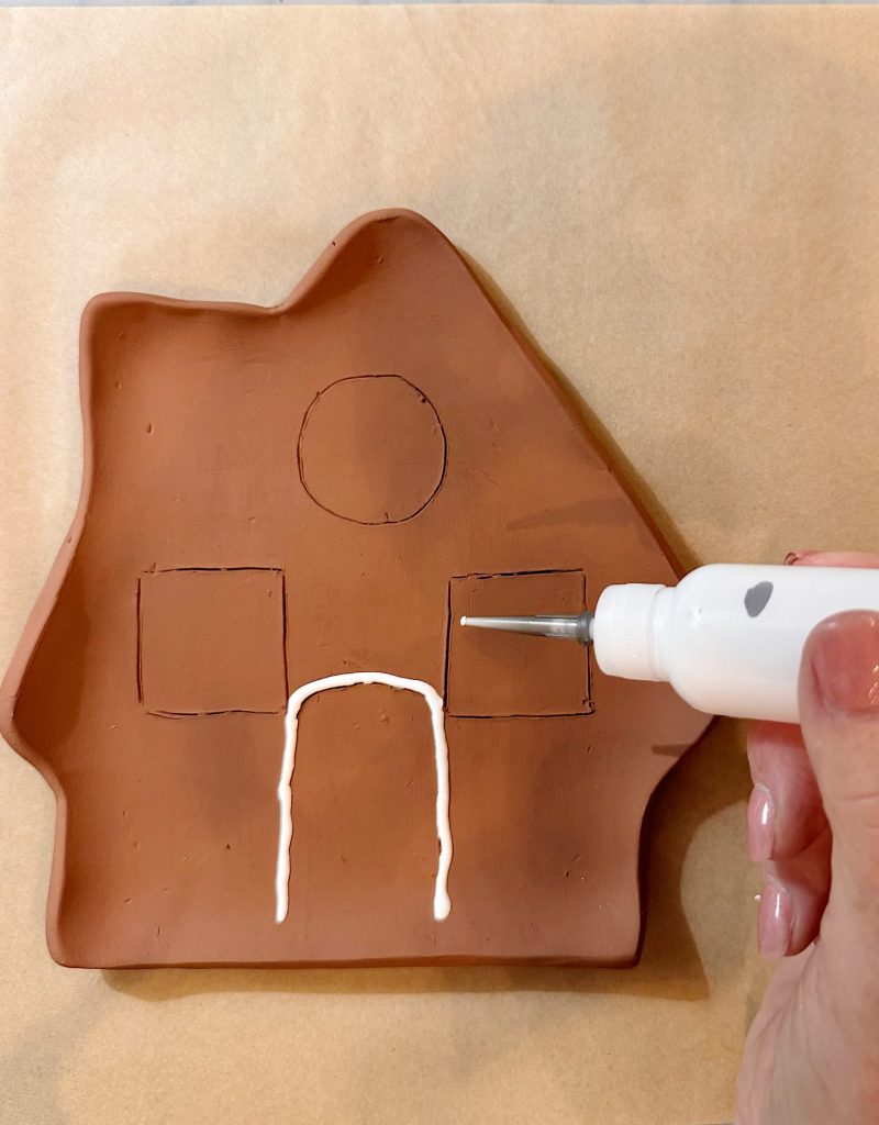 A hand applies white icing to a brown clay tray shaped like a house with outlines for windows, a door, and a circle.