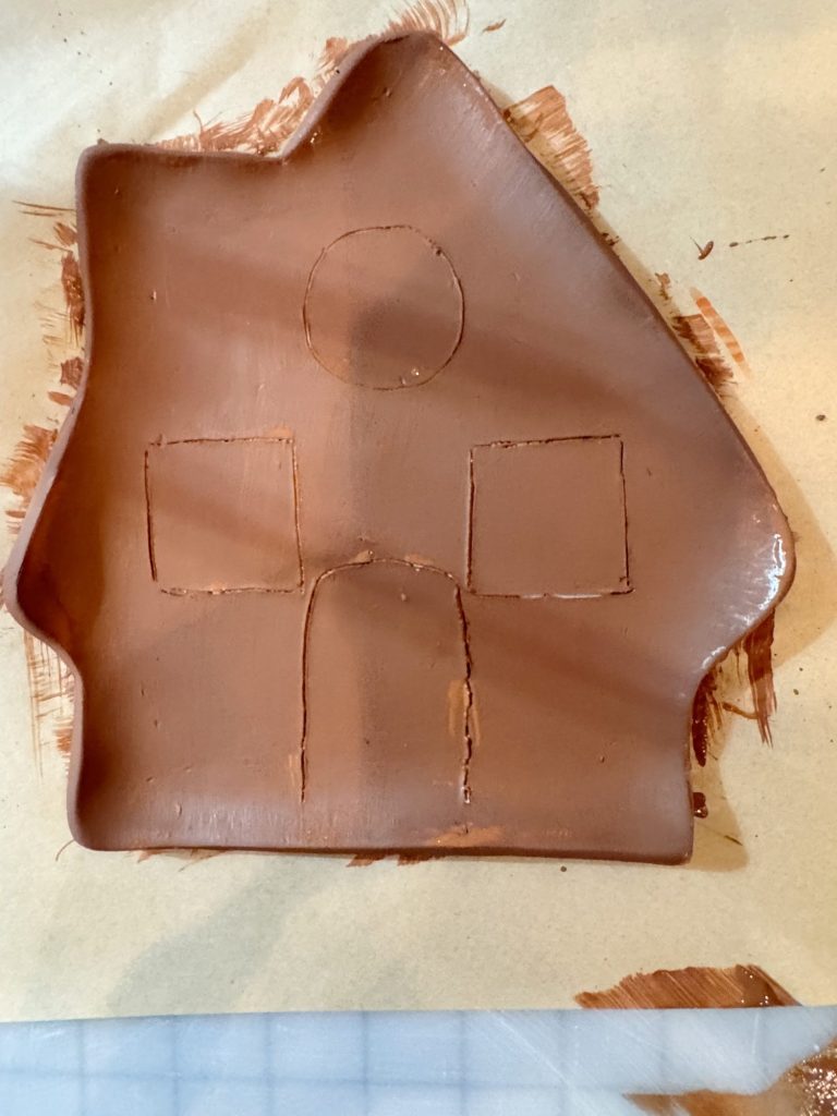 A flat piece of brown clay shaped like a house with square windows, a round window, and a door outlined on its surface.