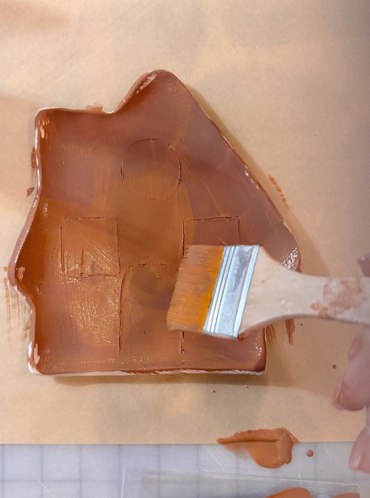 A hand uses a paintbrush to apply brown paint to a clay object resting on a sheet of brown paper.