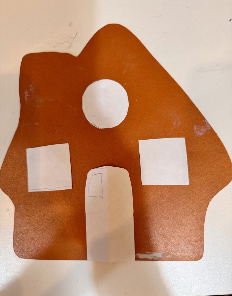 A brown paper cutout shaped like a house with white paper shapes representing two windows, a door, and a round window above the door.