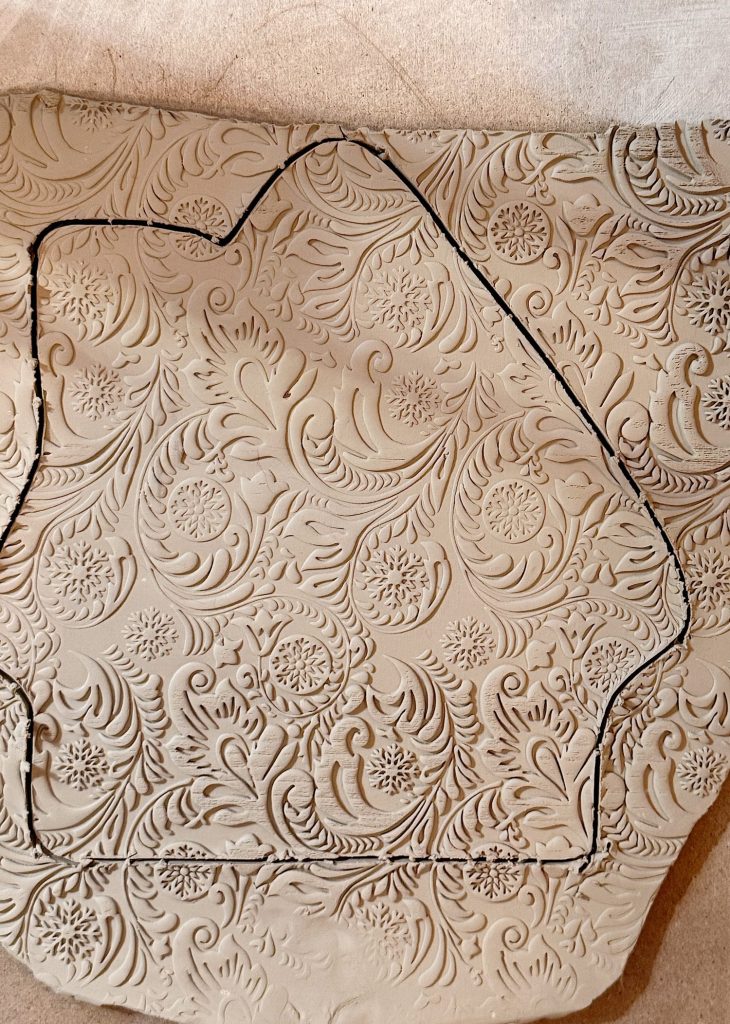 A slab of clay with an ornate, floral and leaf pattern is outlined with a black line in the shape of a house.