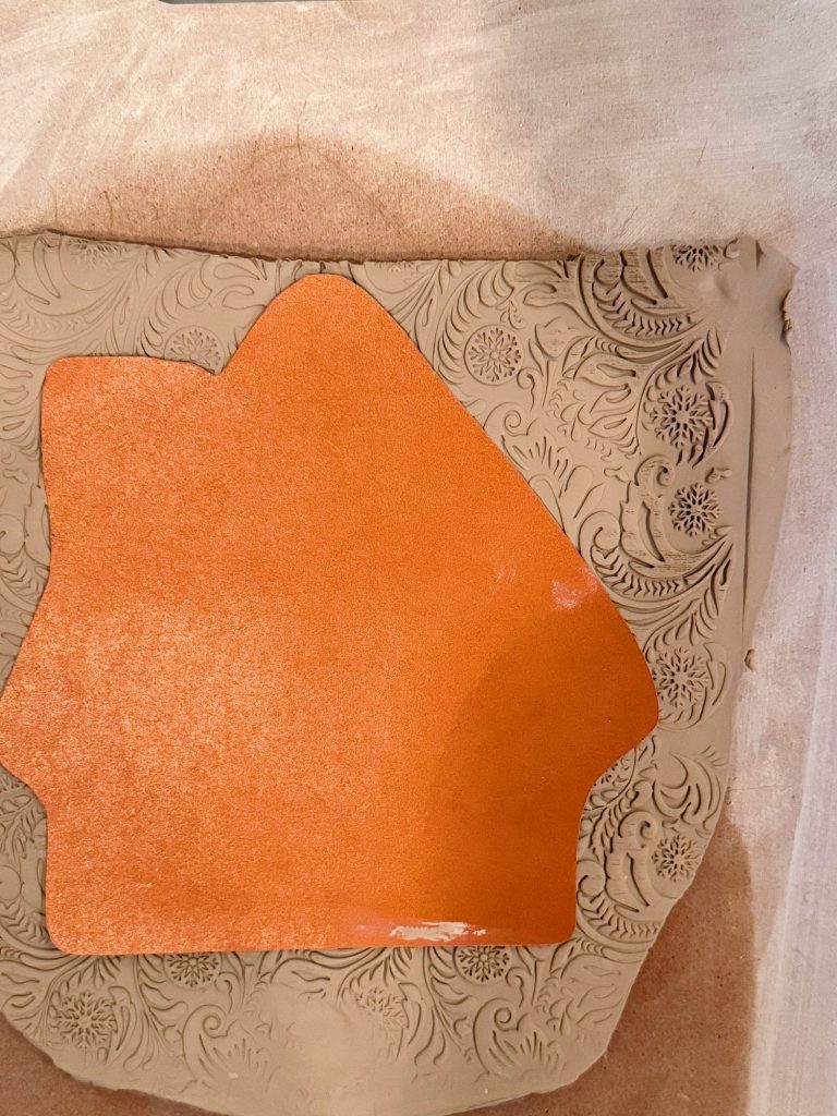 A piece of clay with embossed floral patterns and an orange house-shaped cutout placed on top.