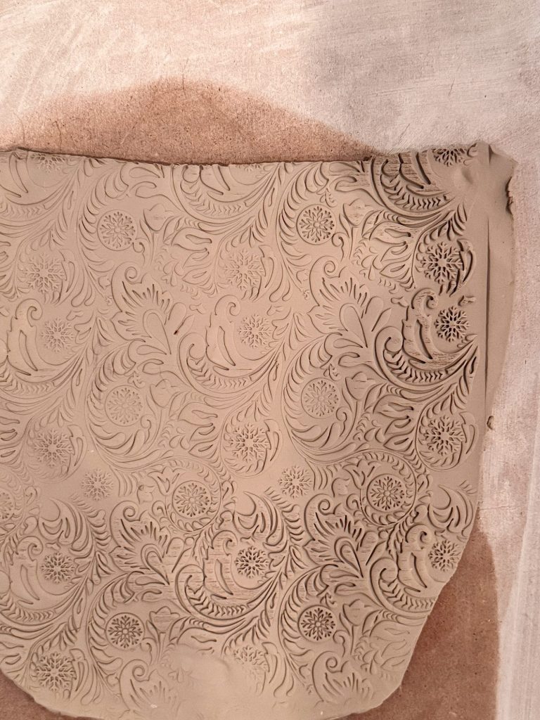 A slab of clay with an intricate floral and leaf pattern embossed on its surface rests on a fabric-covered surface.