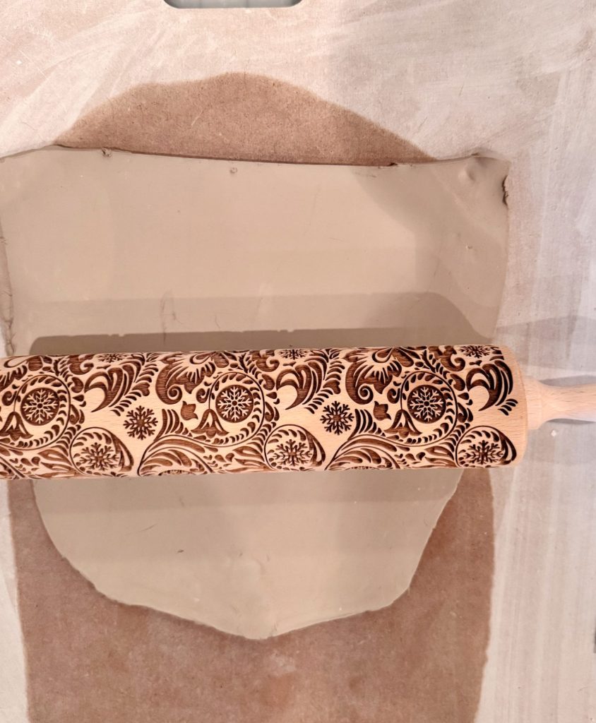 A patterned rolling pin is being used to emboss a decorative design onto a sheet of clay laid out on a fabric surface.