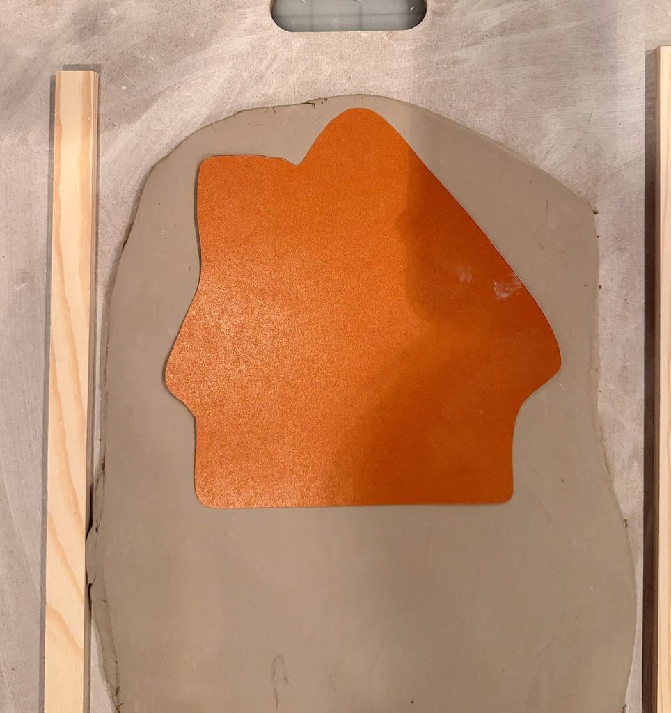 A flat slab of clay with an orange, house-shaped template placed on top, positioned between two wooden guide rails on a work surface.