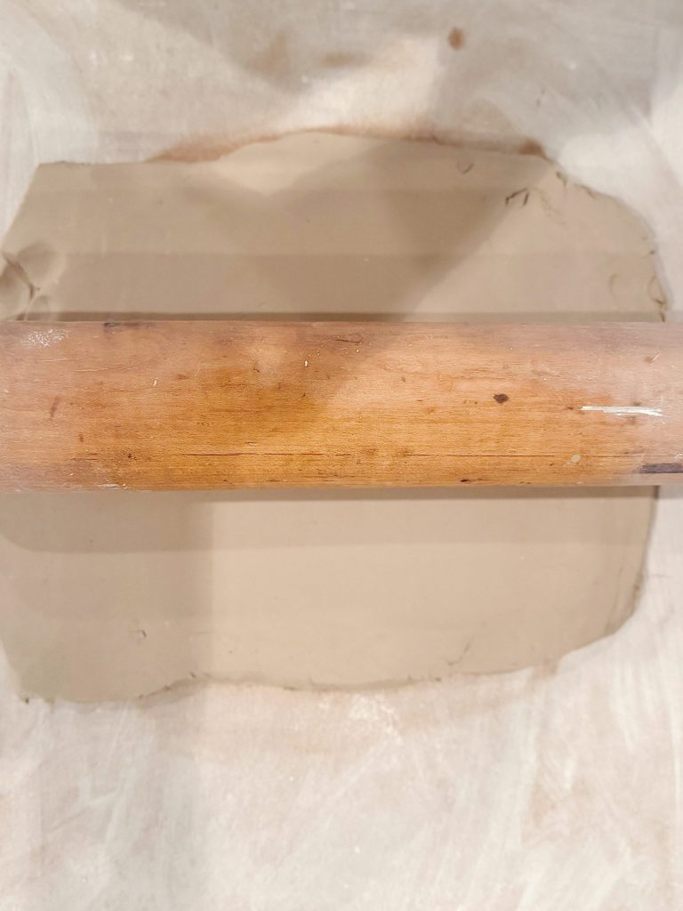 A wooden rolling pin is flattening a slab of light brown clay on a white surface.