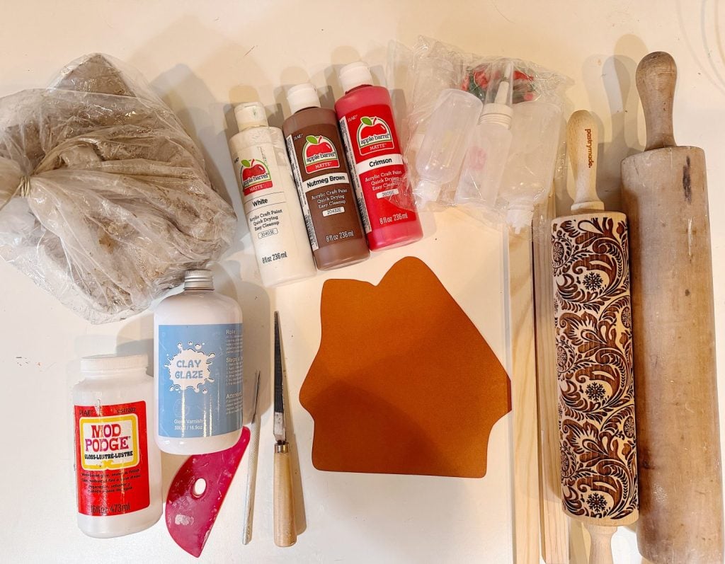 A flat lay of clay, paint bottles, Mod Podge, clay glaze, crafting tools, a plastic scraper, three wooden sticks, and two rolling pins, one with a decorative pattern.