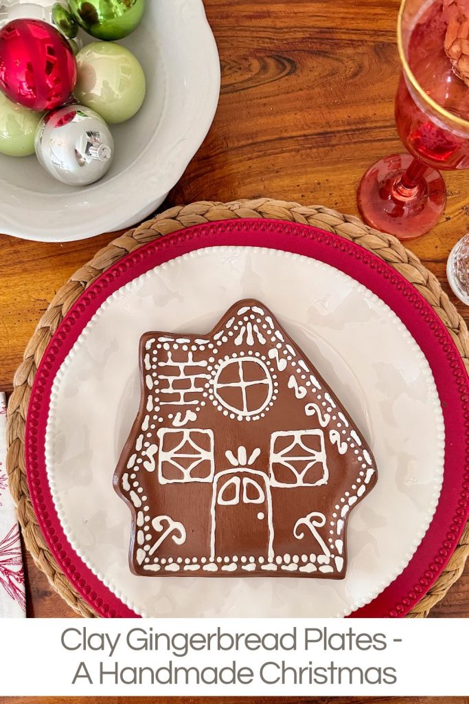 A clay gingerbread house plate with white icing design sits on a white plate with a red border, next to a bowl of ornaments and a glass on a wooden table.