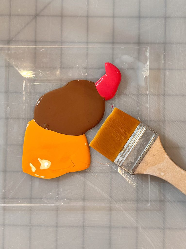 A flat paintbrush rests beside blobs of red, brown, and yellow paint on a transparent surface with a grid background.