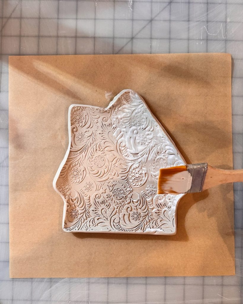 A decorated white clay piece with an ornate floral pattern is being brushed with a clear substance on a brown surface.