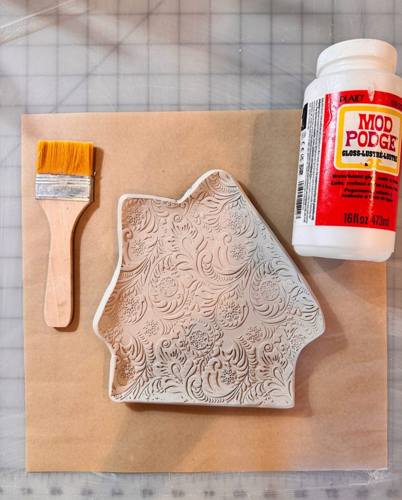 A flat brush, an embossed clay dish, and a jar of Mod Podge gloss sealer are arranged on a brown sheet atop a grid-patterned surface.