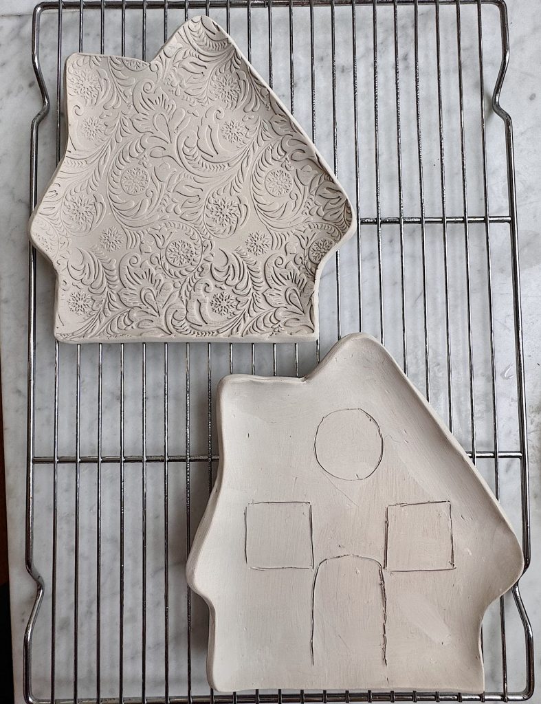 Two house-shaped clay dishes rest on a wire rack; one has a floral embossed pattern, the other has geometric shapes etched into its surface.