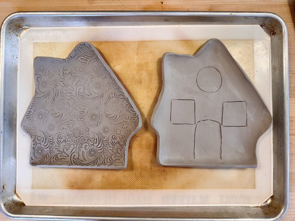 Two house-shaped slabs of clay on a baking tray; the left has a floral pattern, and the right has simple geometric shapes etched into its surface.