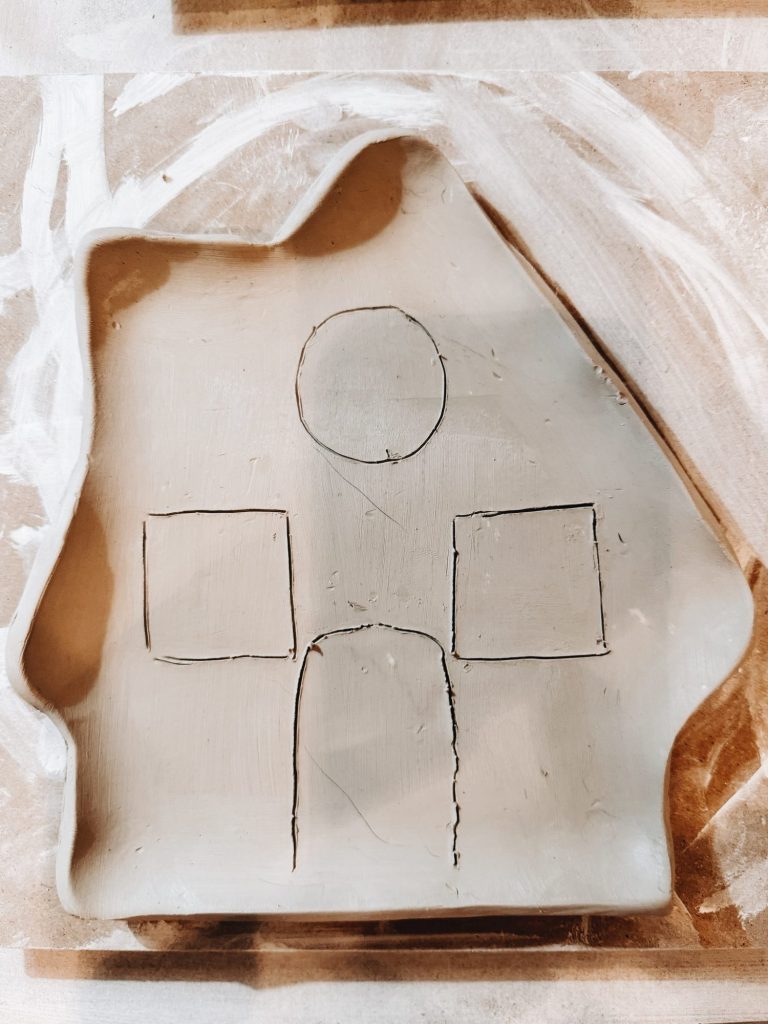 A piece of clay shaped like a house with lines etched to outline a door, two windows, and a circular shape above the door.