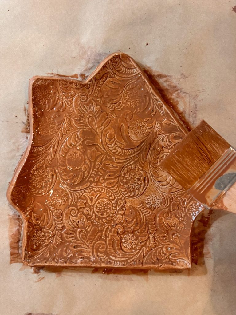 A brown, patterned clay dish in an irregular shape sits on parchment paper, with a brush applying glaze or paint to its surface.