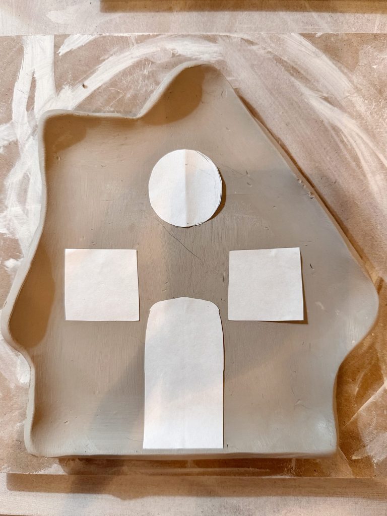 Cut-out paper shapes—one circle, two squares, and one rectangle—are arranged on a clay slab with an irregular, house-like outline.