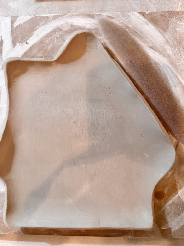 A flat, unfinished clay slab shaped like a house sits on a wooden surface lightly dusted with clay powder.
