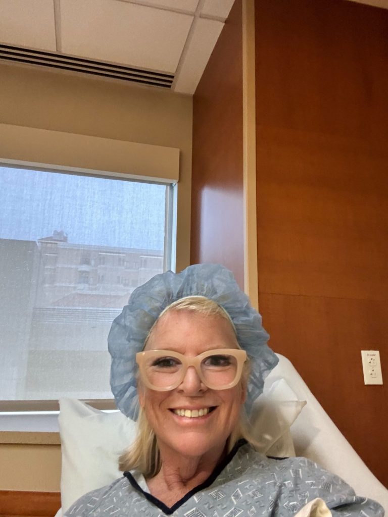 A person wearing a hospital gown, glasses, and a blue surgical cap is smiling while sitting in a hospital bed.
